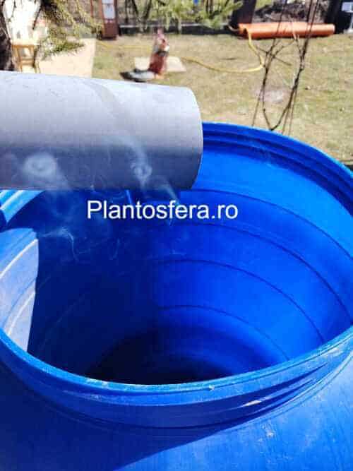 Un rând de butoaie albastre parțial ascunse de plante decorative și arbuști. Culoarea albastră contrastează plăcut cu verdele grădinii, subliniind caracterul ecologic al instalației.